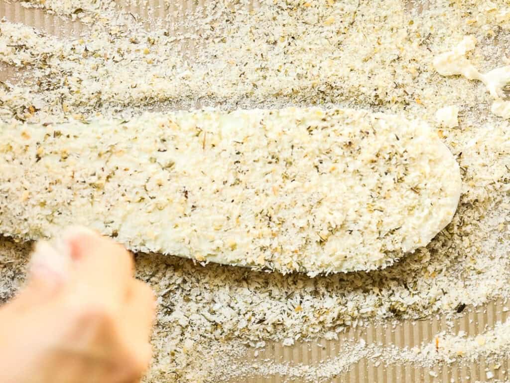 A hand is breading a eggplant fillet with a mixture of breadcrumbs and herbs on a textured surface. The fillet is coated evenly, and breadcrumb flakes are scattered around.